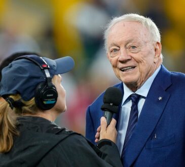 Dallas Cowboys owner Jerry Jones smiles while speaking into a reporter's microphone on the field before a game.