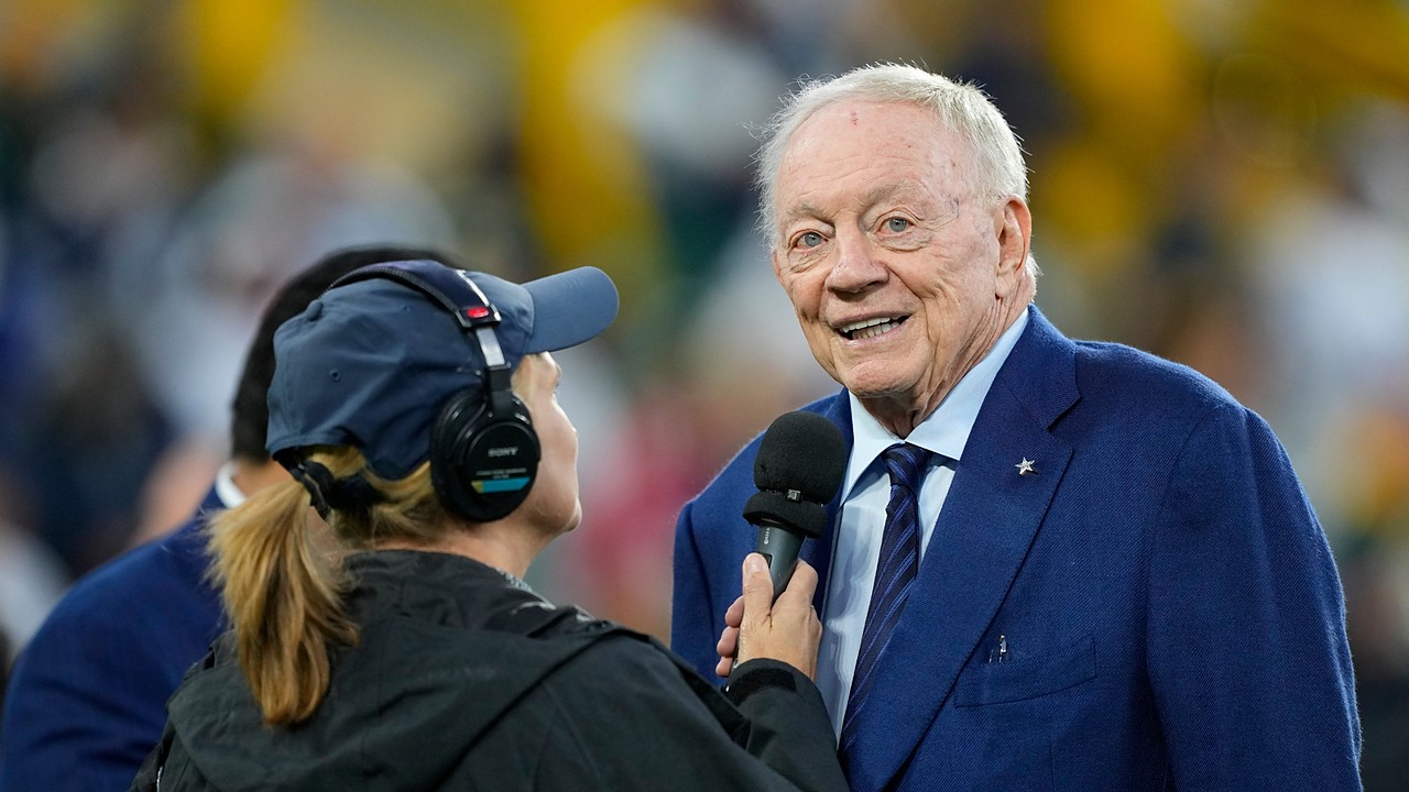 Dallas Cowboys owner Jerry Jones smiles while speaking into a reporter's microphone on the field before a game.