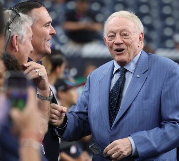 Cowboys owner Jerry Jones smiles and offers a fist bump to fans along the sideline in a blue pinstripe suit.