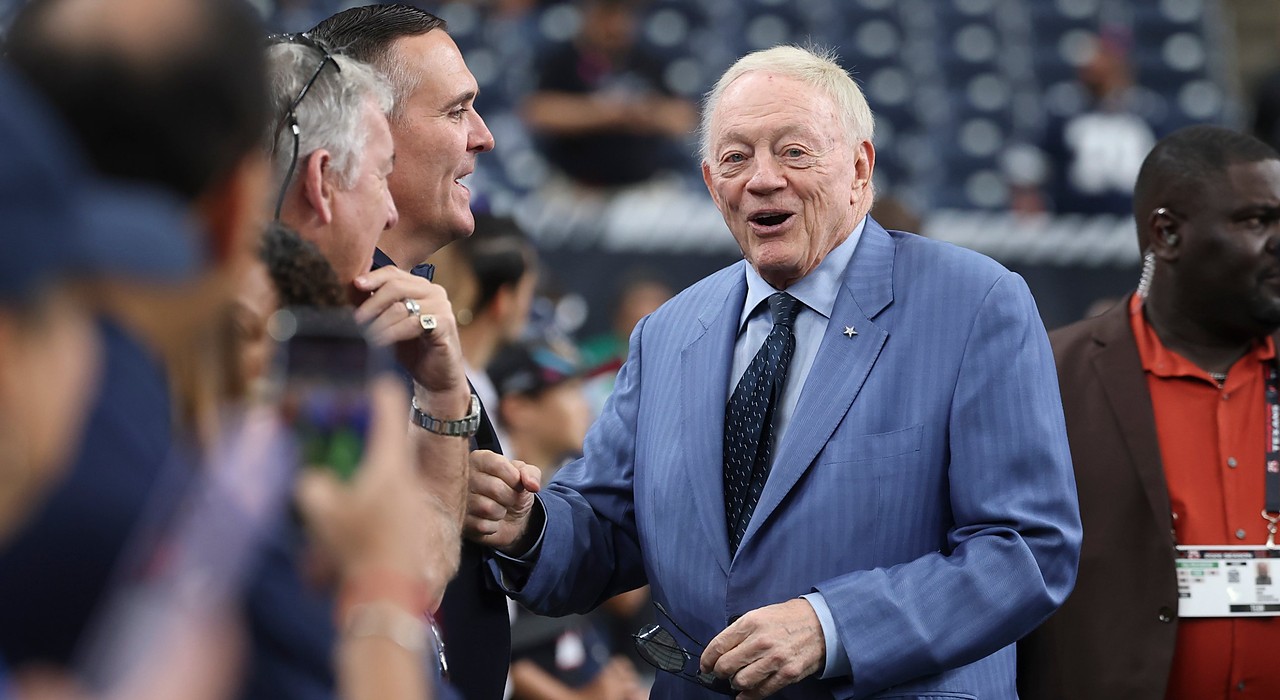 Cowboys owner Jerry Jones smiles and offers a fist bump to fans along the sideline in a blue pinstripe suit.