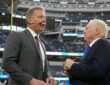 Dallas Cowboys owner Jerry Jones talks with former Cowboys quarterback Troy Aikman, who wears a broadcast headset, on the field.