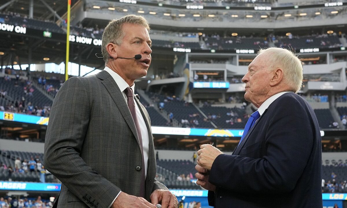 Dallas Cowboys owner Jerry Jones talks with former Cowboys quarterback Troy Aikman, who wears a broadcast headset, on the field.