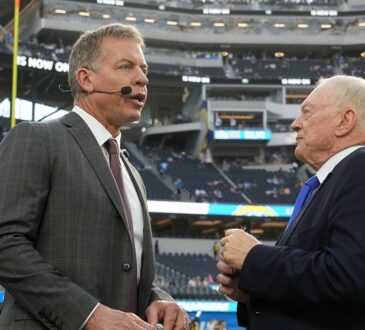 Dallas Cowboys owner Jerry Jones talks with former Cowboys quarterback Troy Aikman, who wears a broadcast headset, on the field.