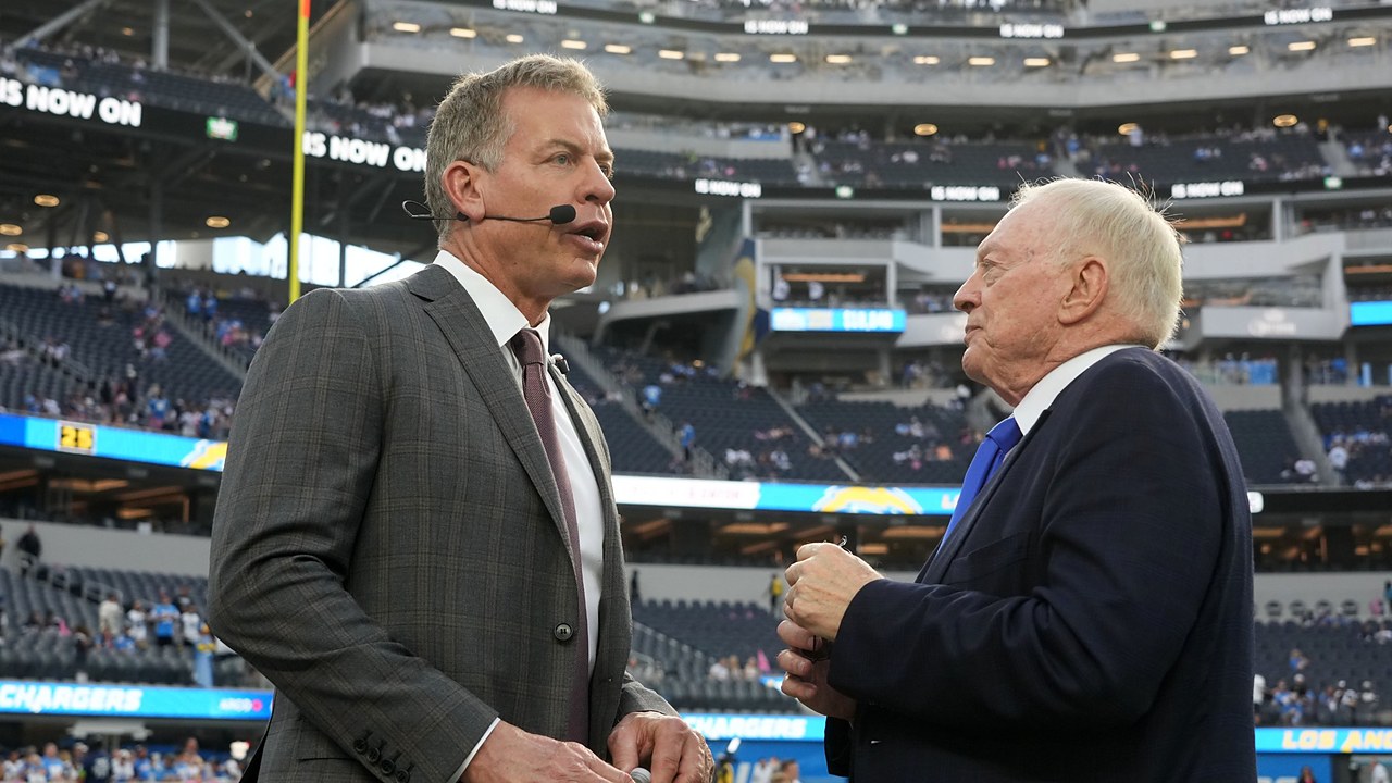 Dallas Cowboys owner Jerry Jones talks with former Cowboys quarterback Troy Aikman, who wears a broadcast headset, on the field.