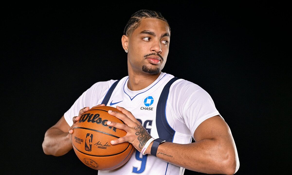Dallas Mavericks guard Jett Howard holds a basketball at his chest and glances to his left against a black backdrop.