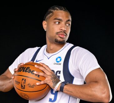 Dallas Mavericks guard Jett Howard holds a basketball at his chest and glances to his left against a black backdrop.