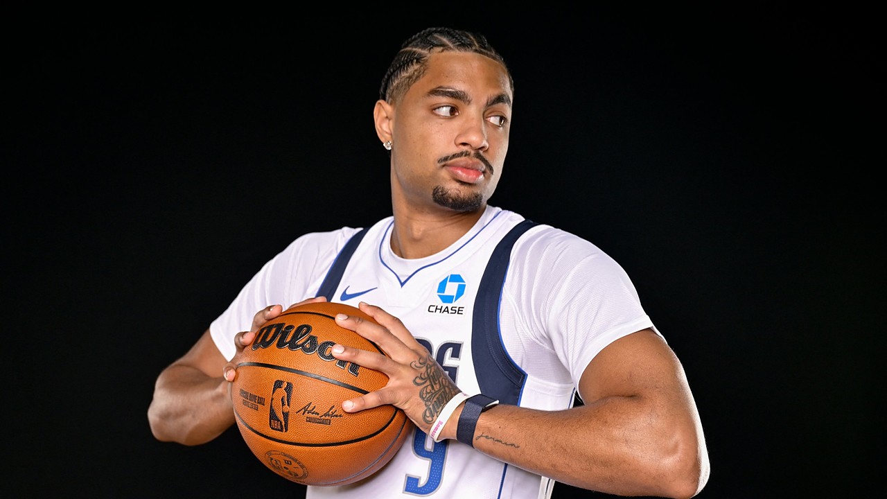 Dallas Mavericks guard Jett Howard holds a basketball at his chest and glances to his left against a black backdrop.
