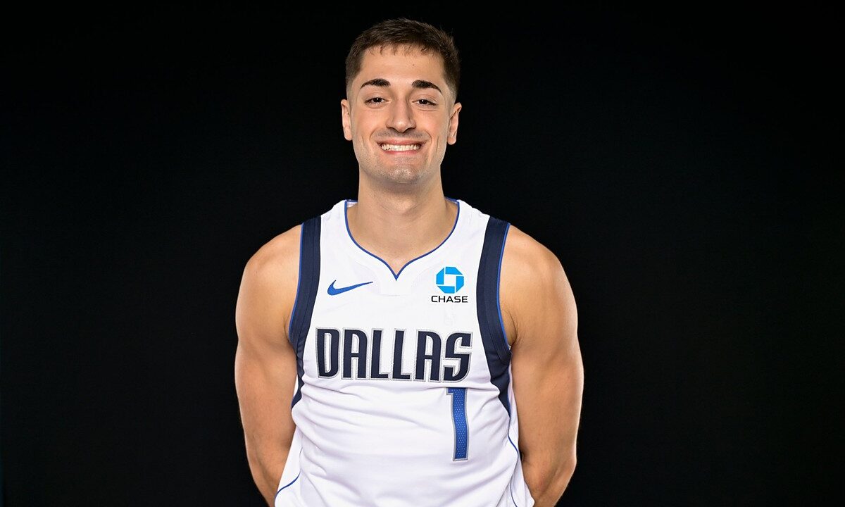 Dallas Mavericks guard John Poulakidas smiles with his hands behind his back in a white Dallas No. 1 jersey.
