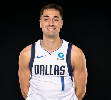 Dallas Mavericks guard John Poulakidas smiles with his hands behind his back in a white Dallas No. 1 jersey.