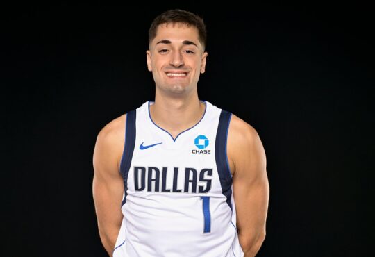 Dallas Mavericks guard John Poulakidas smiles with his hands behind his back in a white Dallas No. 1 jersey.
