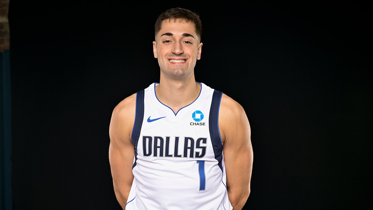 Dallas Mavericks guard John Poulakidas smiles with his hands behind his back in a white Dallas No. 1 jersey.