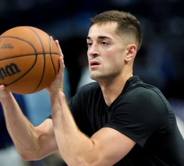 Dallas Mavericks guard John Poulakidas, in a black T-shirt, holds a basketball in shooting position during warmups.