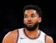New York Knicks center Karl-Anthony Towns glances to the side in a white jersey with orange and blue trim.