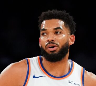 New York Knicks center Karl-Anthony Towns glances to the side in a white jersey with orange and blue trim.