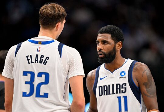 Dallas Mavericks guard Kyrie Irving talks with forward Cooper Flagg, who has his back to the camera, during a break in play.