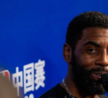 Mavericks guard Kyrie Irving smiles while speaking into a microphone in front of an NBA China Games backdrop.