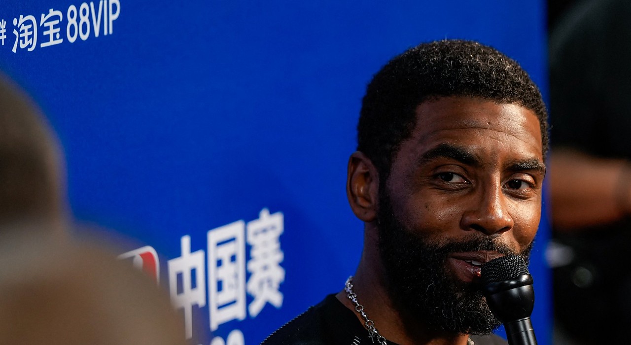 Mavericks guard Kyrie Irving smiles while speaking into a microphone in front of an NBA China Games backdrop.