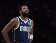 Dallas Mavericks guard Kyrie Irving looks up with his hands on his hips in a navy MAVS jersey against a dark background.