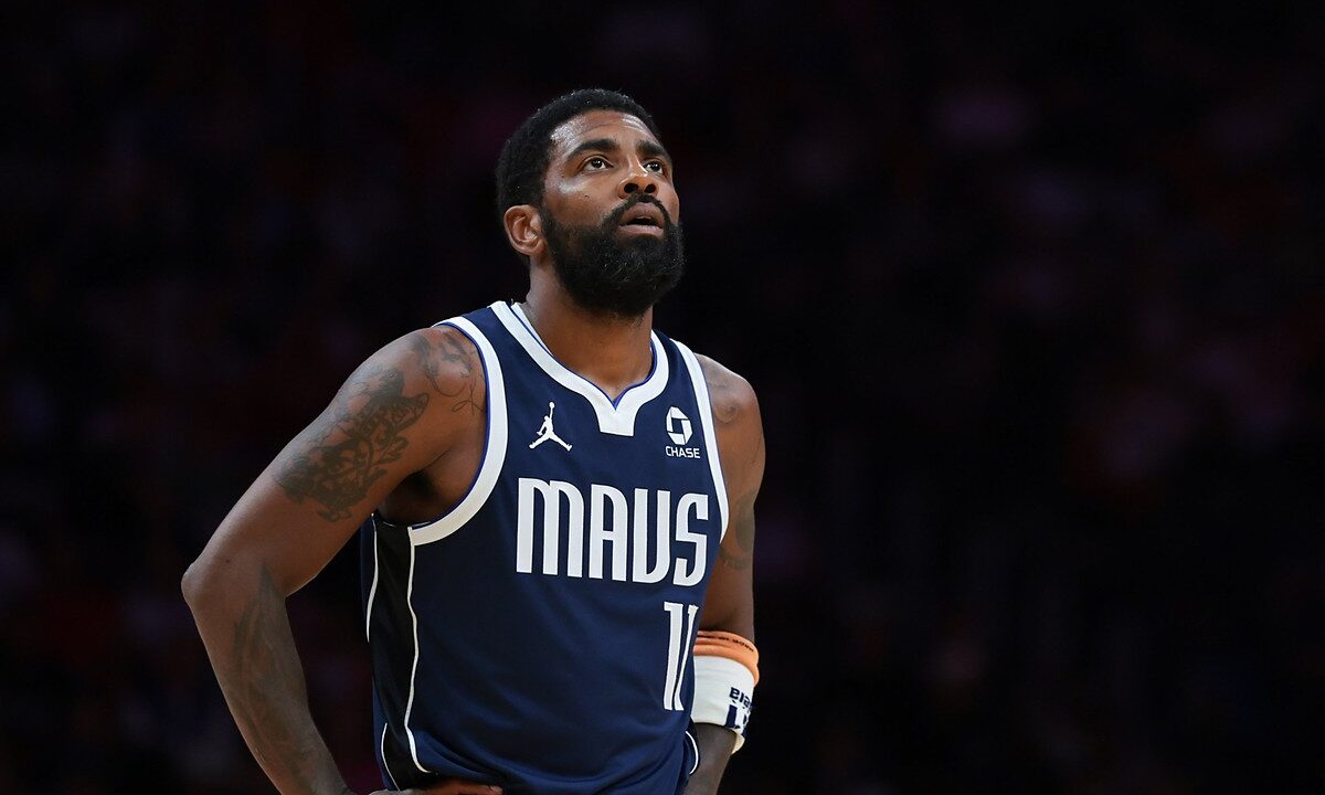 Dallas Mavericks guard Kyrie Irving looks up with his hands on his hips in a navy MAVS jersey against a dark background.