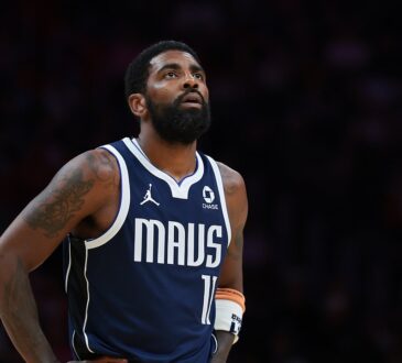 Dallas Mavericks guard Kyrie Irving looks up with his hands on his hips in a navy MAVS jersey against a dark background.