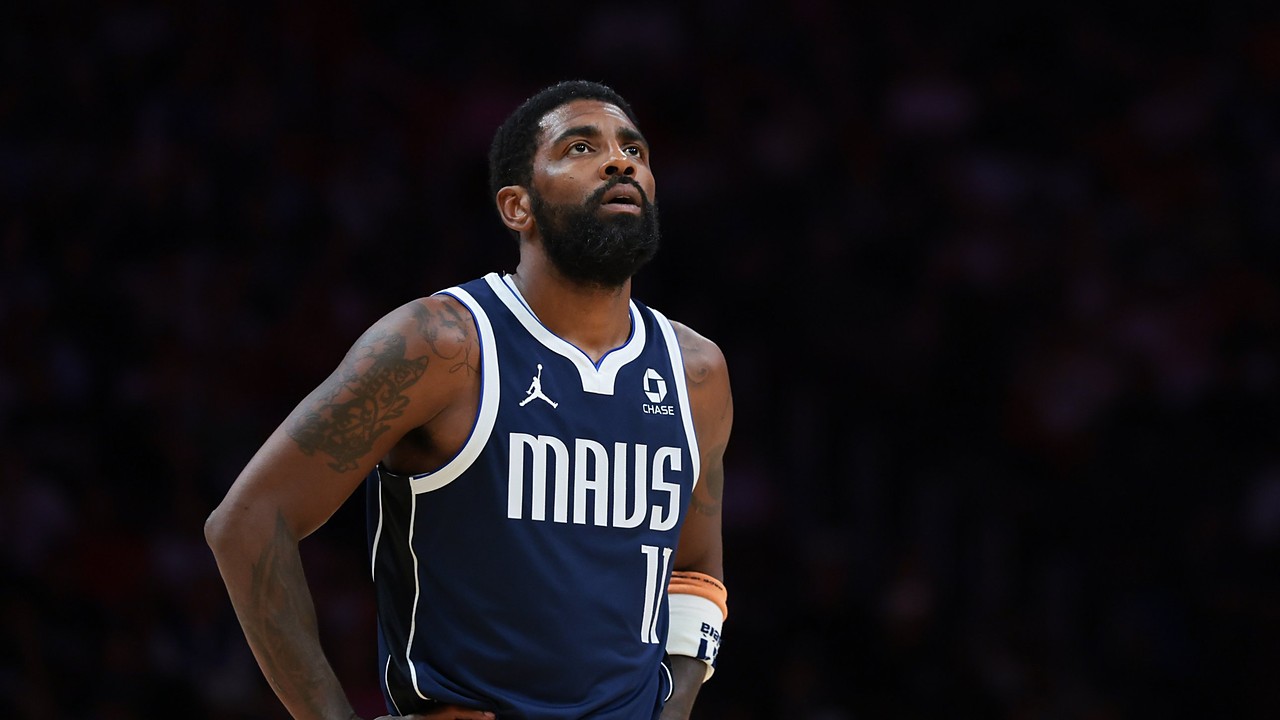 Dallas Mavericks guard Kyrie Irving looks up with his hands on his hips in a navy MAVS jersey against a dark background.