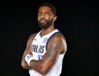 Mavericks guard Kyrie Irving poses with his arms crossed in a white Dallas jersey against a black backdrop.