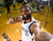 Mavericks guard Kyrie Irving grins and points at himself with one arm stretched toward the camera as media work behind him.