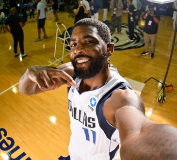Mavericks guard Kyrie Irving grins and points at himself with one arm stretched toward the camera as media work behind him.
