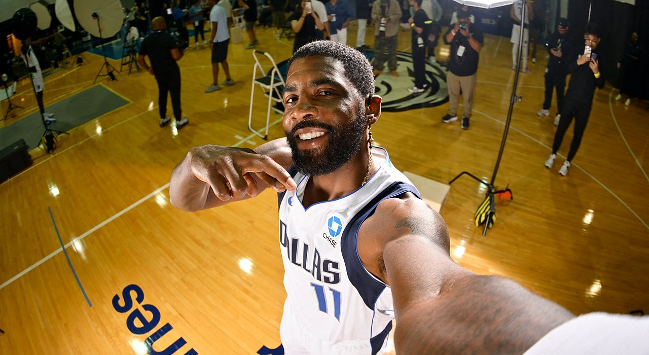 Mavericks guard Kyrie Irving grins and points at himself with one arm stretched toward the camera as media work behind him.