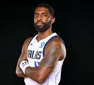 Mavericks guard Kyrie Irving poses with his arms crossed in a white Dallas jersey against a black backdrop.