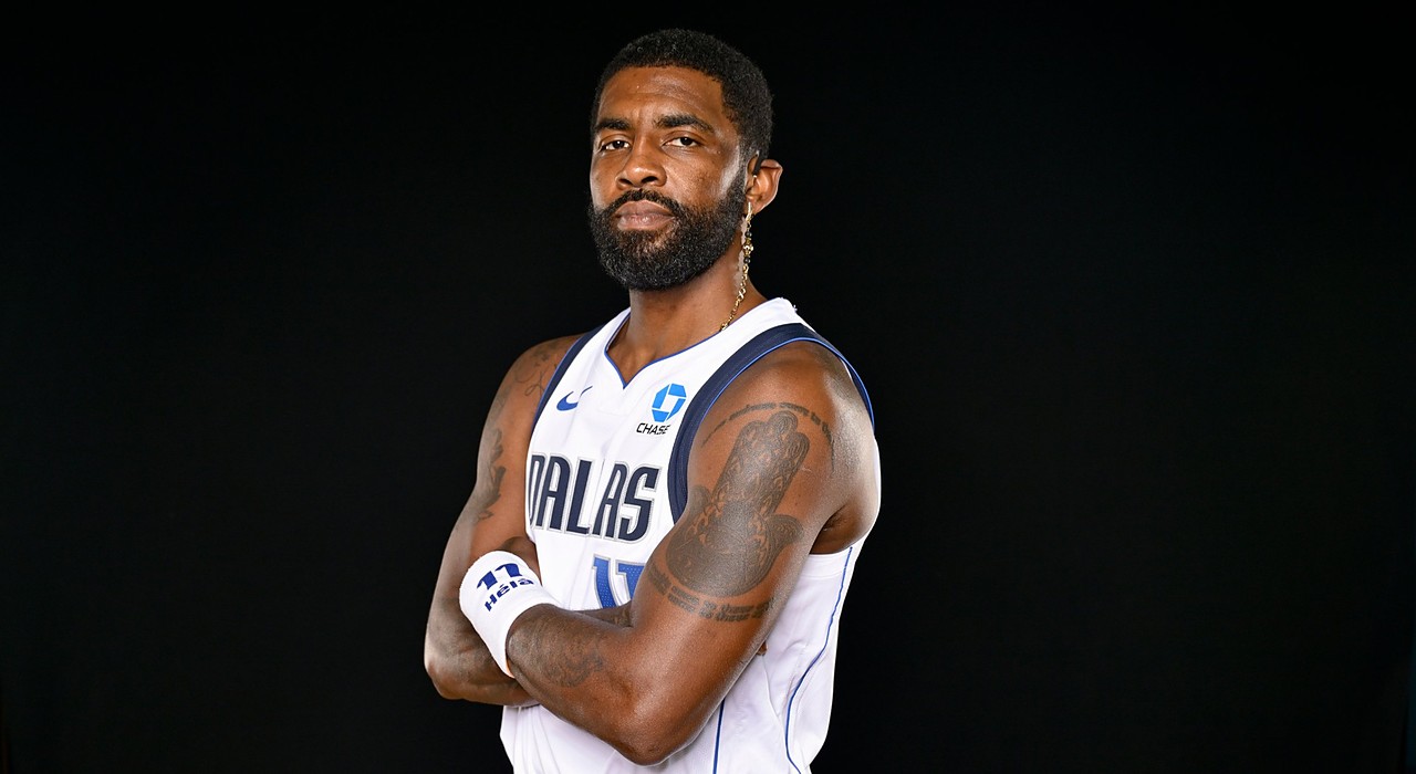 Mavericks guard Kyrie Irving poses with his arms crossed in a white Dallas jersey against a black backdrop.