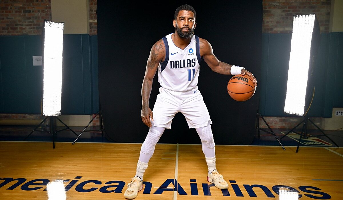 Mavericks guard Kyrie Irving poses in a low dribbling stance in his white home uniform on a studio court.