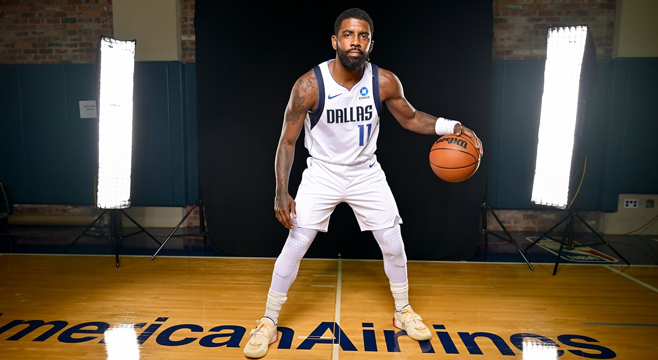 Mavericks guard Kyrie Irving poses in a low dribbling stance in his white home uniform on a studio court.