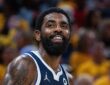 Dallas Mavericks guard Kyrie Irving smiles on the court in a navy Mavericks jersey with a gold-clad crowd blurred behind him.