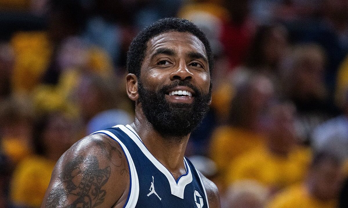Dallas Mavericks guard Kyrie Irving smiles on the court in a navy Mavericks jersey with a gold-clad crowd blurred behind him.