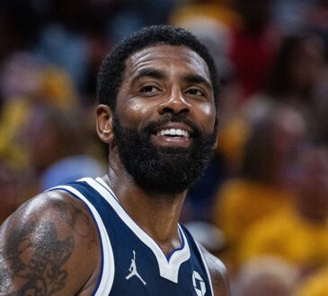 Dallas Mavericks guard Kyrie Irving smiles on the court in a navy Mavericks jersey with a gold-clad crowd blurred behind him.