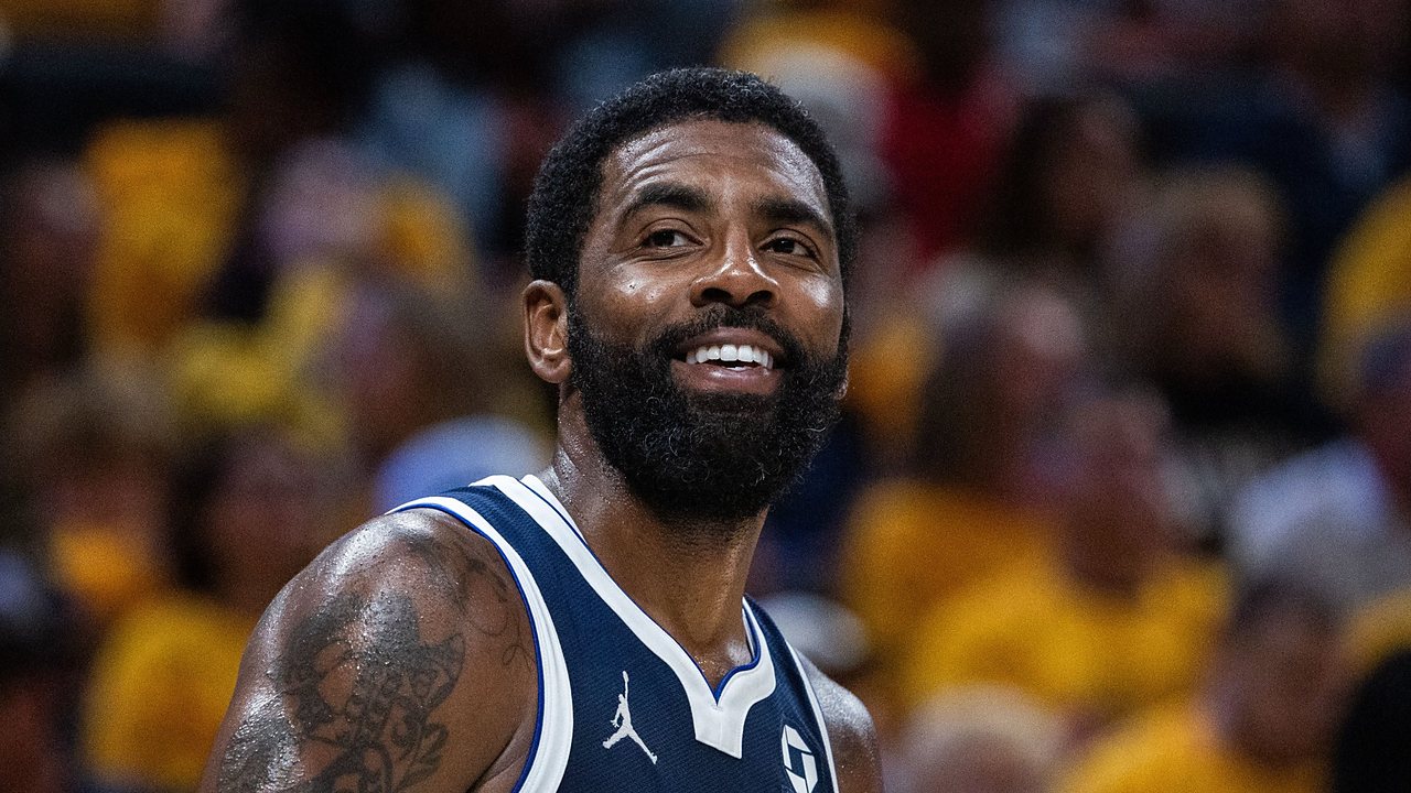 Dallas Mavericks guard Kyrie Irving smiles on the court in a navy Mavericks jersey with a gold-clad crowd blurred behind him.