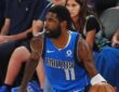 Mavericks guard Kyrie Irving dribbles near the sideline in a blue Dallas jersey as courtside fans look on.