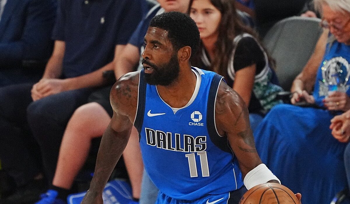 Mavericks guard Kyrie Irving dribbles near the sideline in a blue Dallas jersey as courtside fans look on.