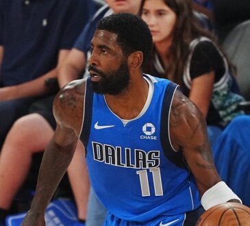 Mavericks guard Kyrie Irving dribbles near the sideline in a blue Dallas jersey as courtside fans look on.