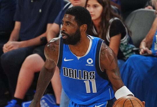 Mavericks guard Kyrie Irving dribbles near the sideline in a blue Dallas jersey as courtside fans look on.