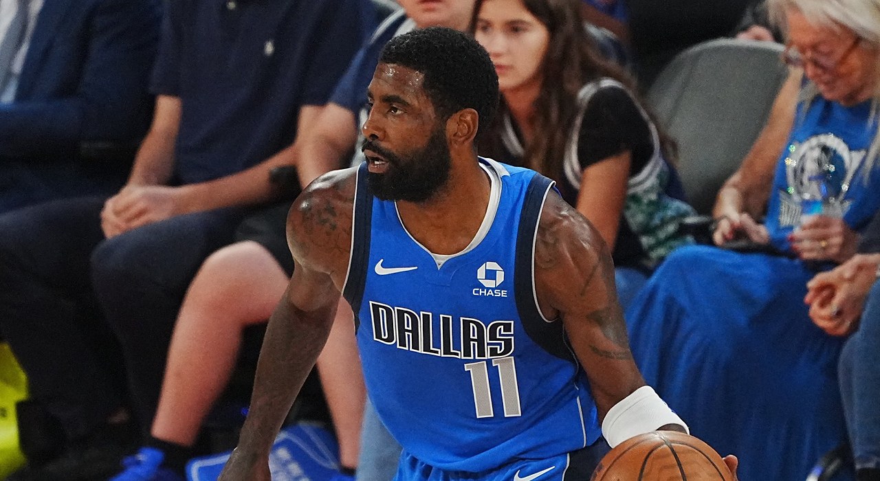 Mavericks guard Kyrie Irving dribbles near the sideline in a blue Dallas jersey as courtside fans look on.