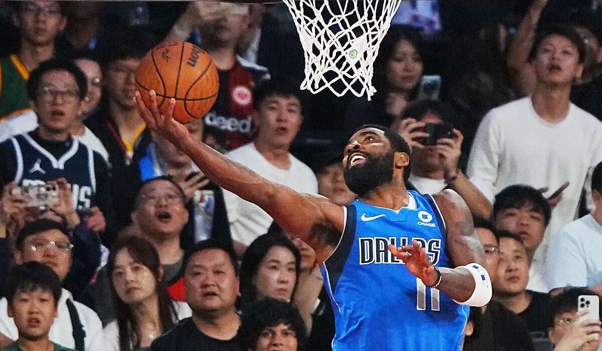 Mavericks guard Kyrie Irving extends for a reverse layup under the rim as fans behind the basket react.