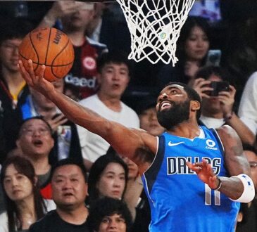 Mavericks guard Kyrie Irving extends for a reverse layup under the rim as fans behind the basket react.