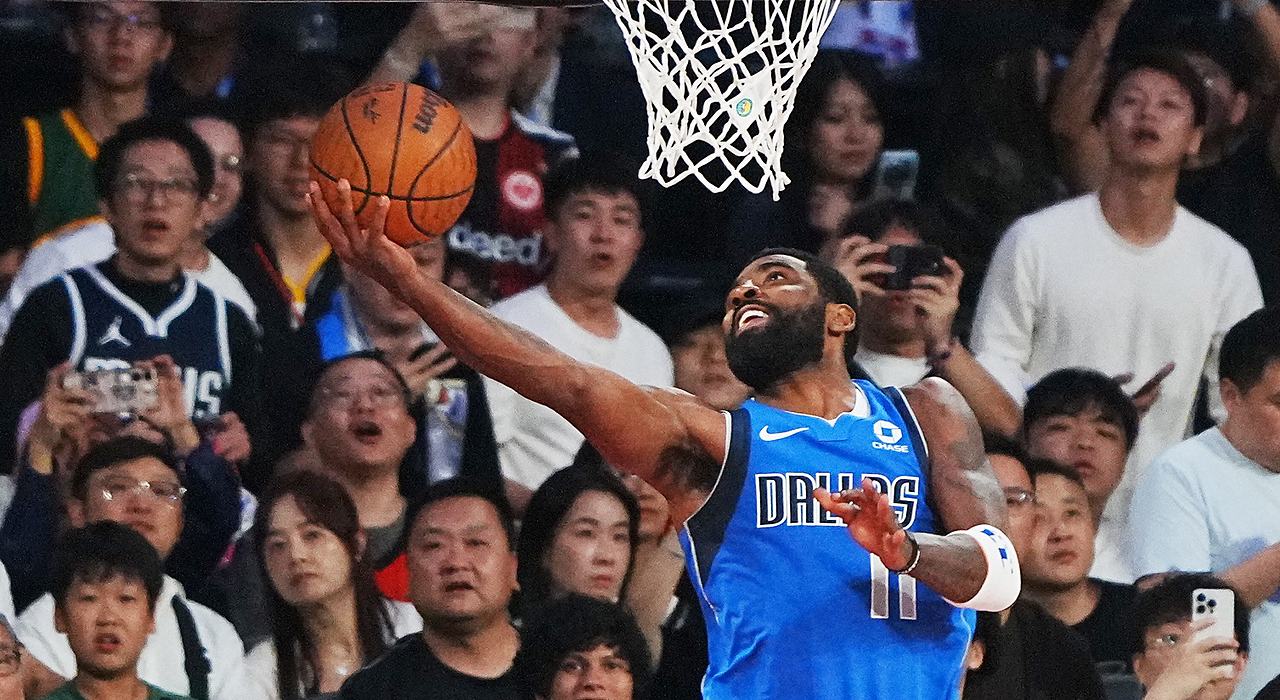 Mavericks guard Kyrie Irving extends for a reverse layup under the rim as fans behind the basket react.