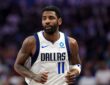 Dallas Mavericks guard Kyrie Irving jogs up the court in a white home jersey with the crowd blurred behind him.