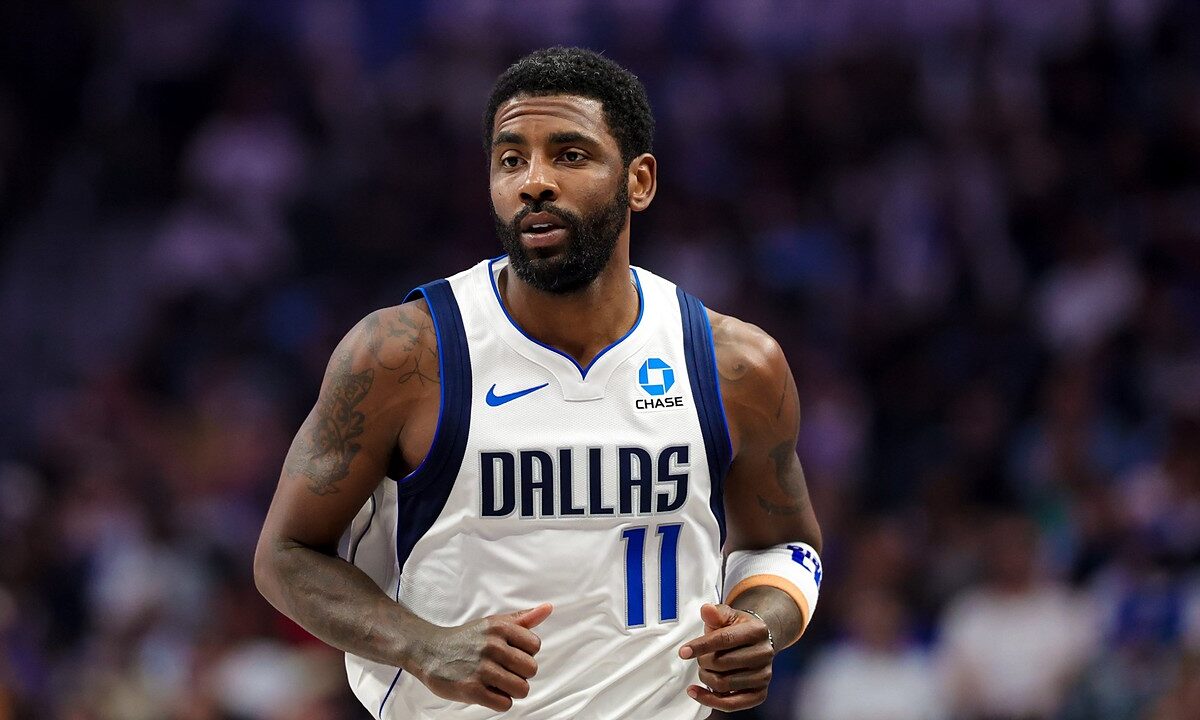 Dallas Mavericks guard Kyrie Irving jogs up the court in a white home jersey with the crowd blurred behind him.