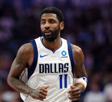 Dallas Mavericks guard Kyrie Irving jogs up the court in a white home jersey with the crowd blurred behind him.