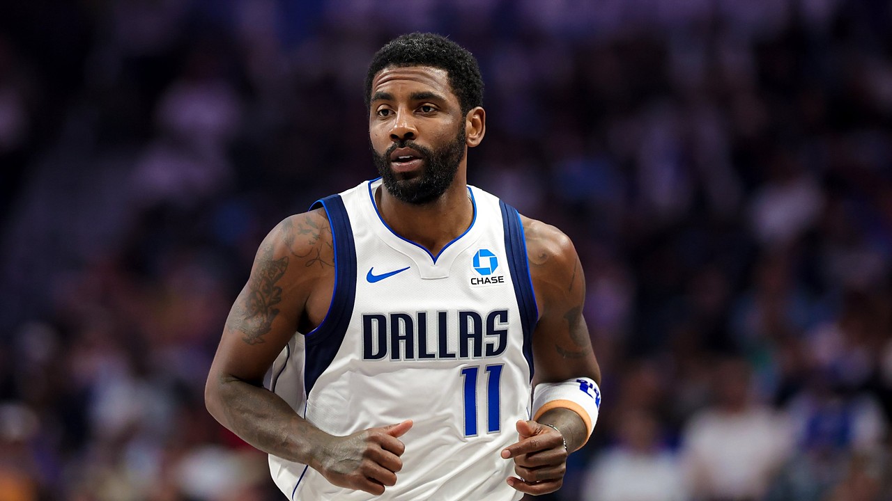 Dallas Mavericks guard Kyrie Irving jogs up the court in a white home jersey with the crowd blurred behind him.