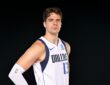 Dallas Mavericks forward Tarik Biberović stands with a hand on his hip in a white Dallas jersey against a black backdrop.